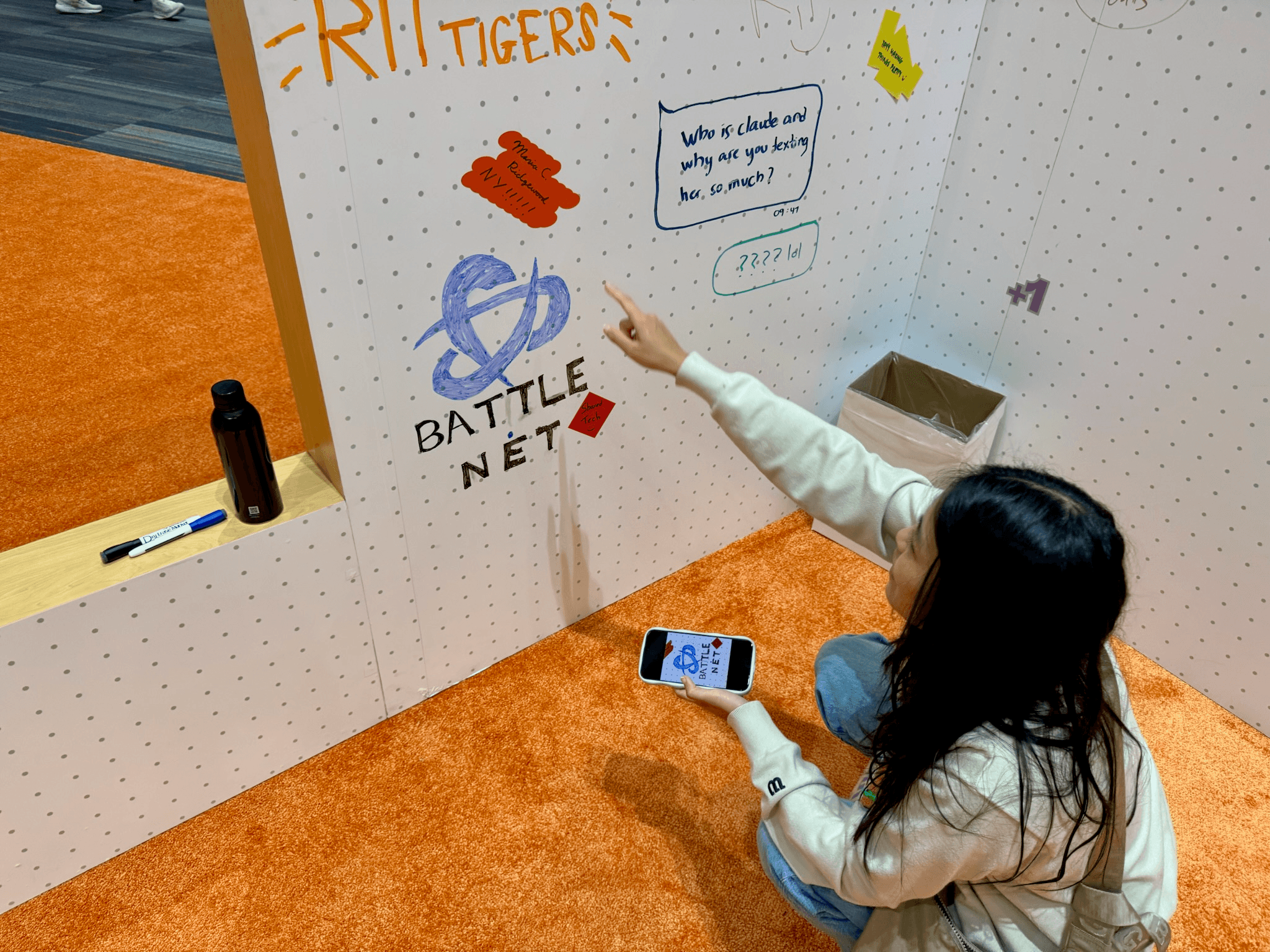 A person pointing at a hand-drawn Battle.net logo on a communal whiteboard wall covered in doodles and notes