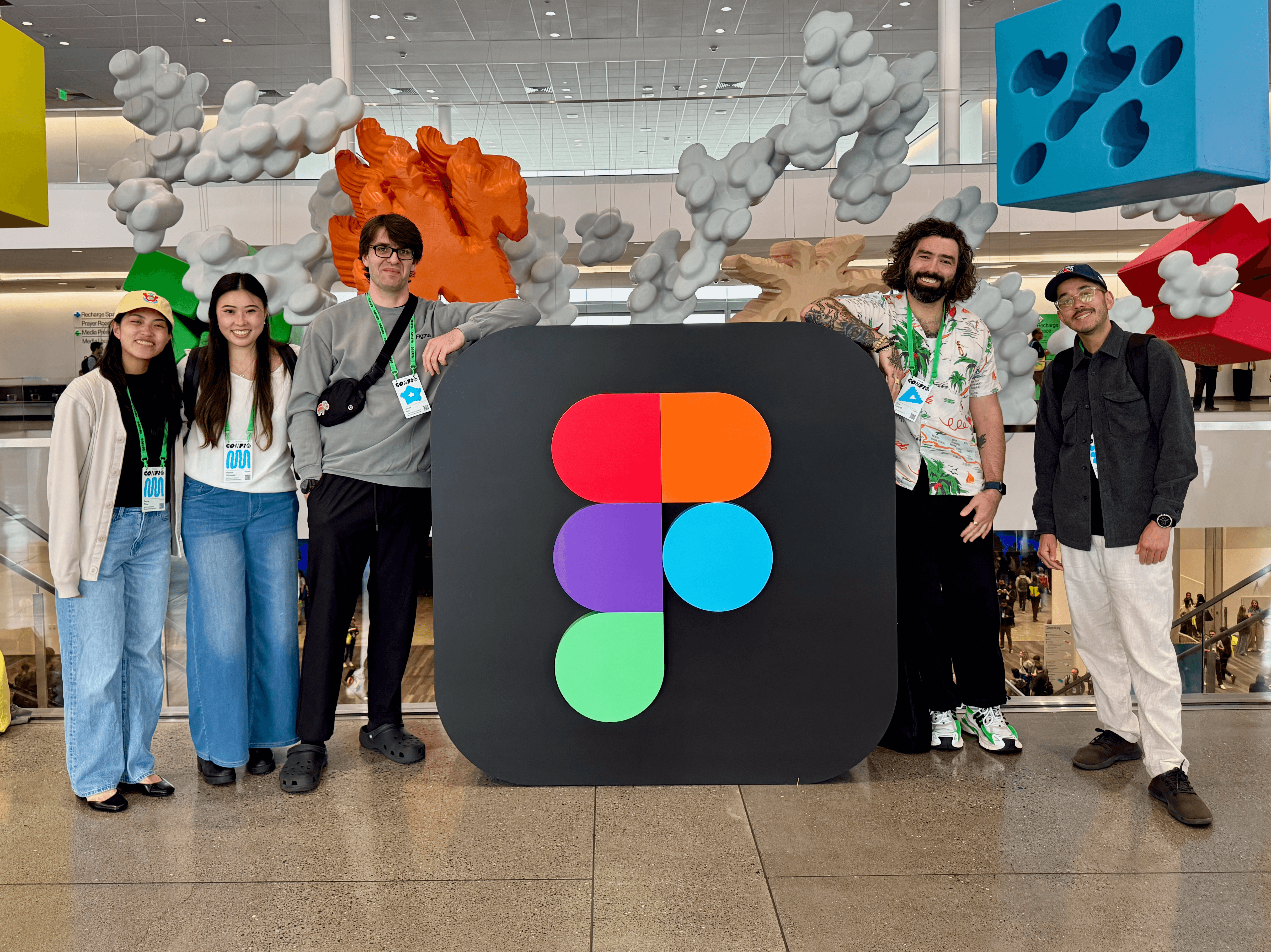 Five conference attendees posing in front of a large 3D Figma logo, with abstract cloud-like sculptures hanging overhead