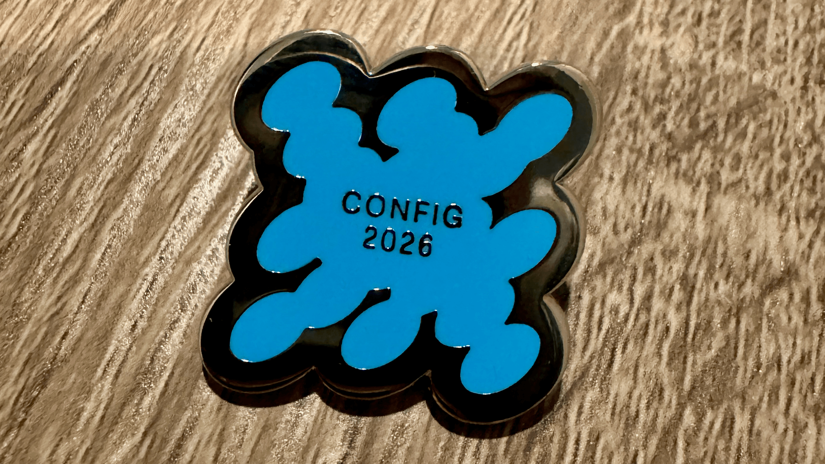 A blue enamel pin shaped like a paint splatter, reading 'Config 2026'