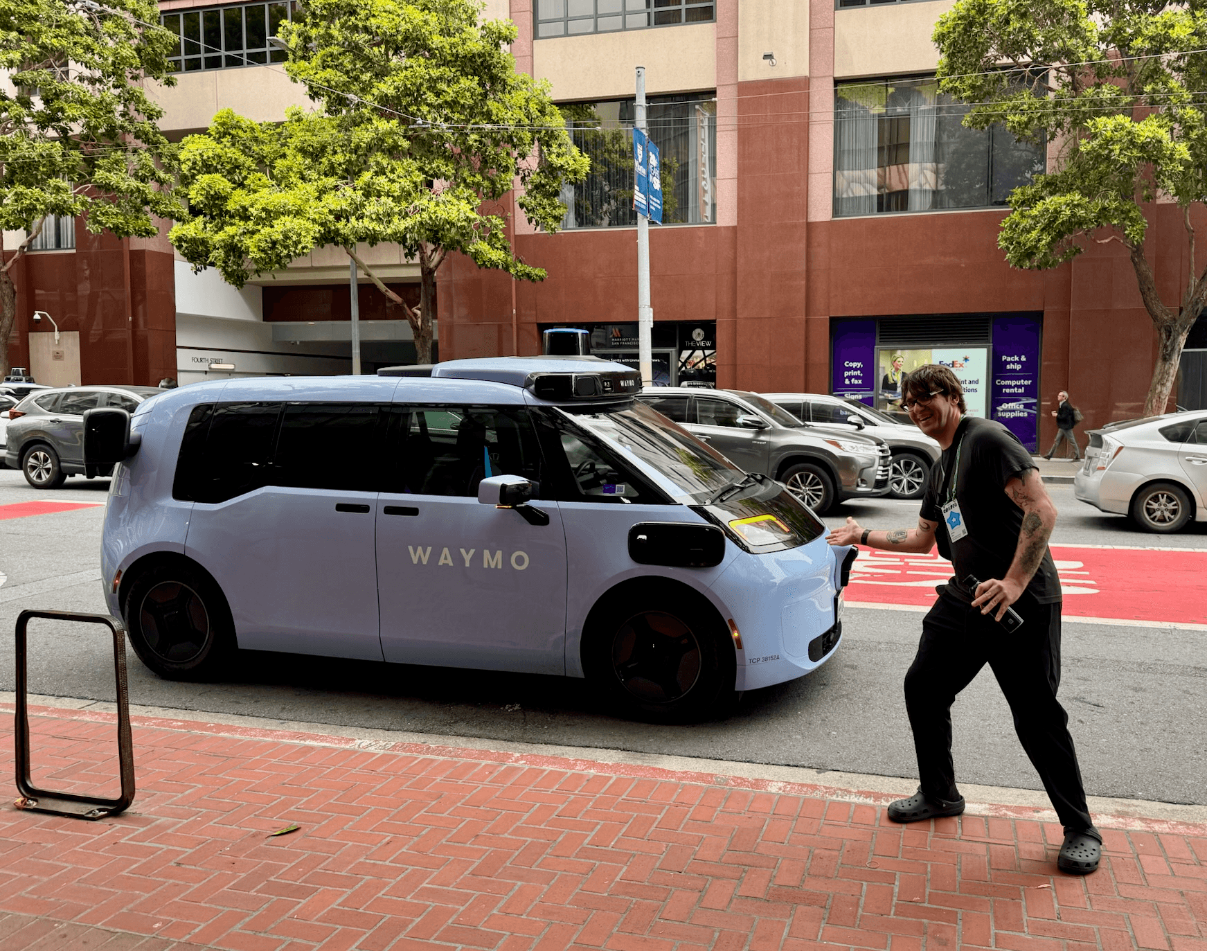 A pale blue Waymo Ojai autonomous vehicle parked on a San Francisco street, with a smiling person walking towards it
