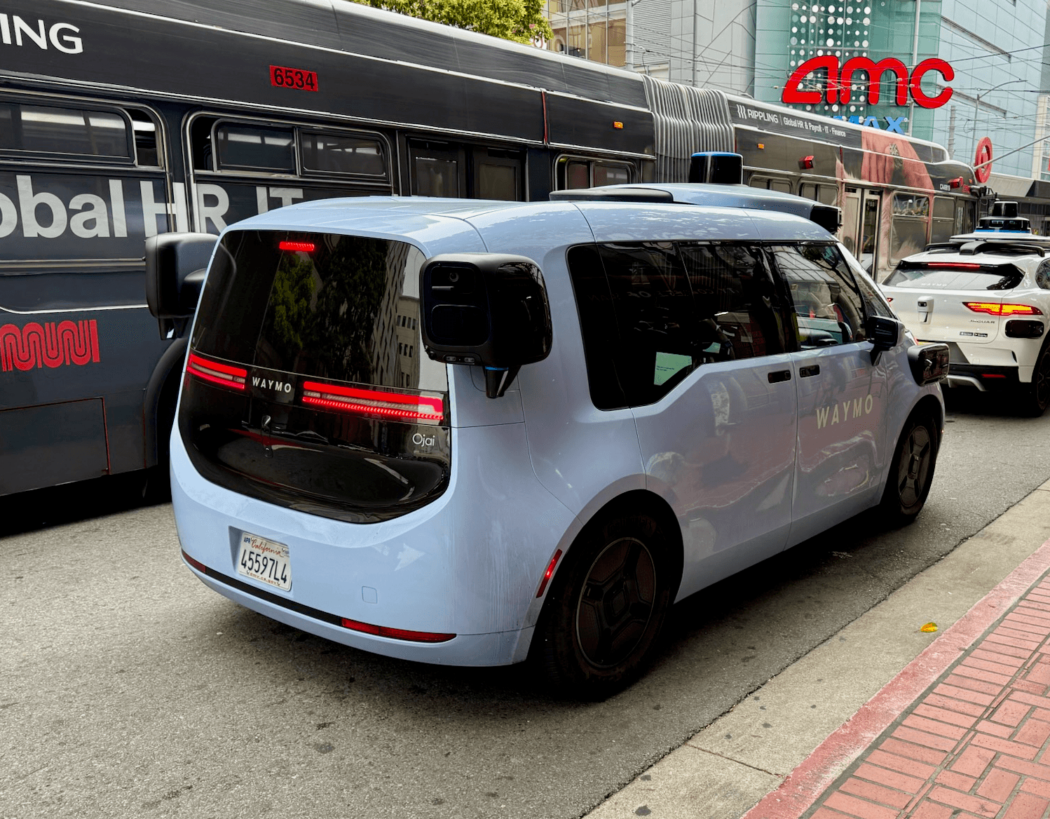 The rear of a pale blue Waymo Ojai autonomous vehicle in traffic, showing its 'Ojai' badge and roof-mounted sensors